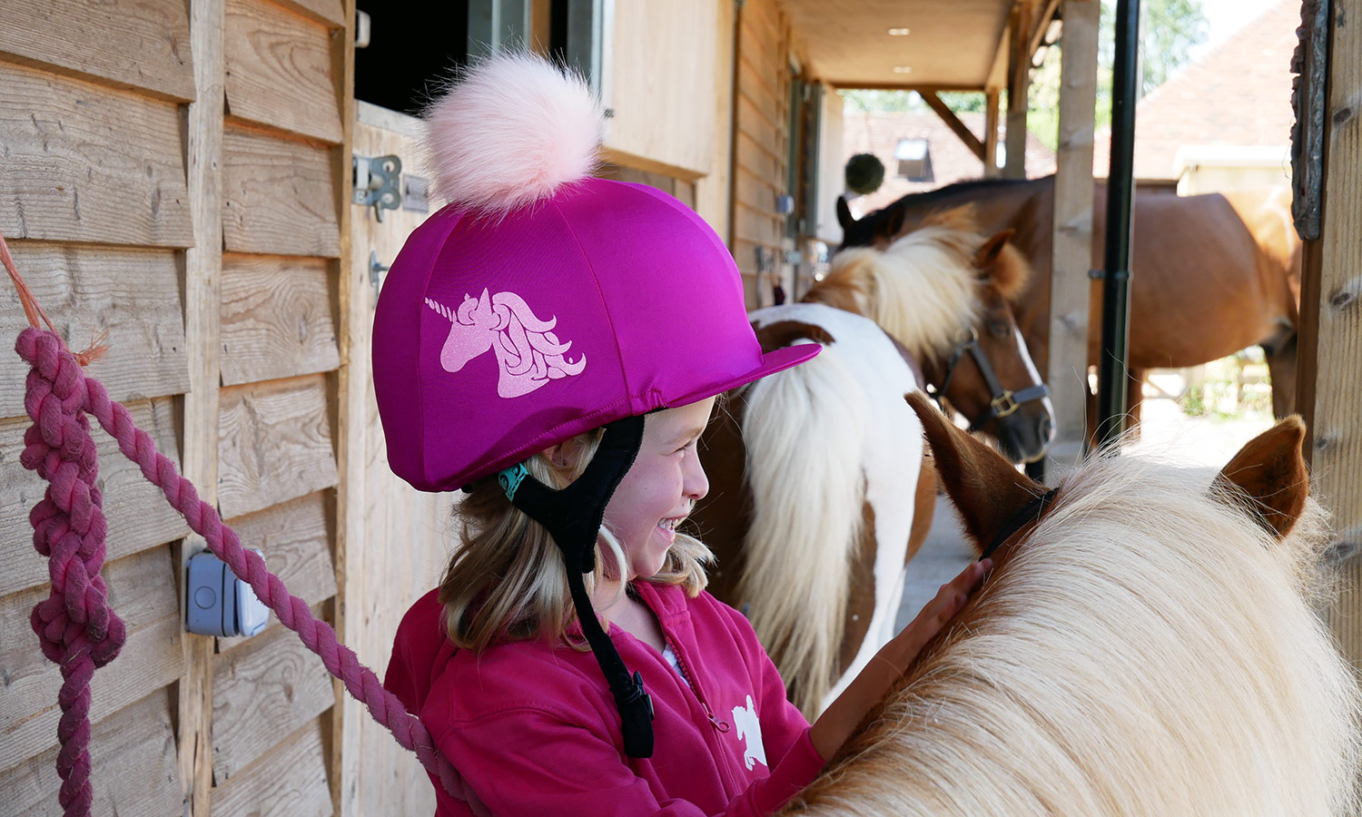 A picture of a young girl, a horse riding beginner wearing a brightly coloured riding hat standing alongside a pony. Picture is by luvponies.com